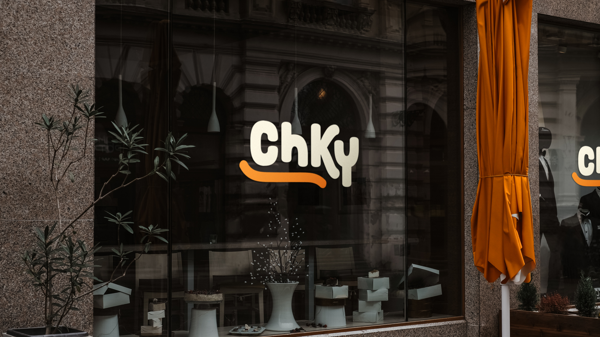 Canva Freelancers 25 Canvajoy Unlimited Graphic Design Storefront with a large window displaying the ChKY logo in bold letters. Inside, various items are showcased on white display stands, reflecting a design subscription flair. An orange umbrella is folded near the entrance of the stone building.